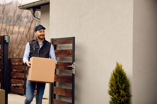 Leende budbärare levererar ett paket till en privatbostad genom grind, som del av bud- och transporttjänst.