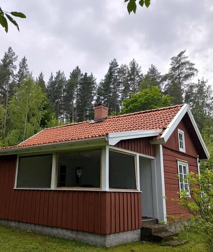 Röd enplansvilla med tegeltak och inglasad altan, omgiven av grönska och skog i bakgrunden.