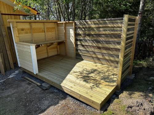Halvfärdig uteplats eller förråd i trä med sittbänk, byggt på trall, omgiven av skog.