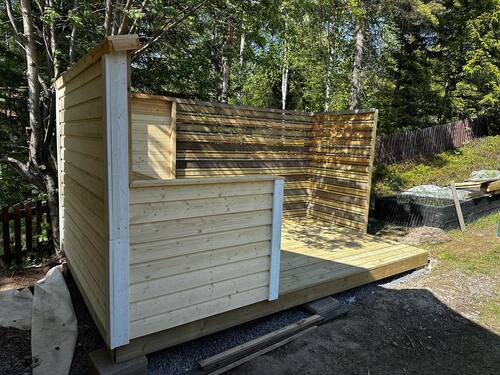 Träkonstruktion under uppbyggnad i trädgård, med väggar i stående panel och öppen fram- och taksida.