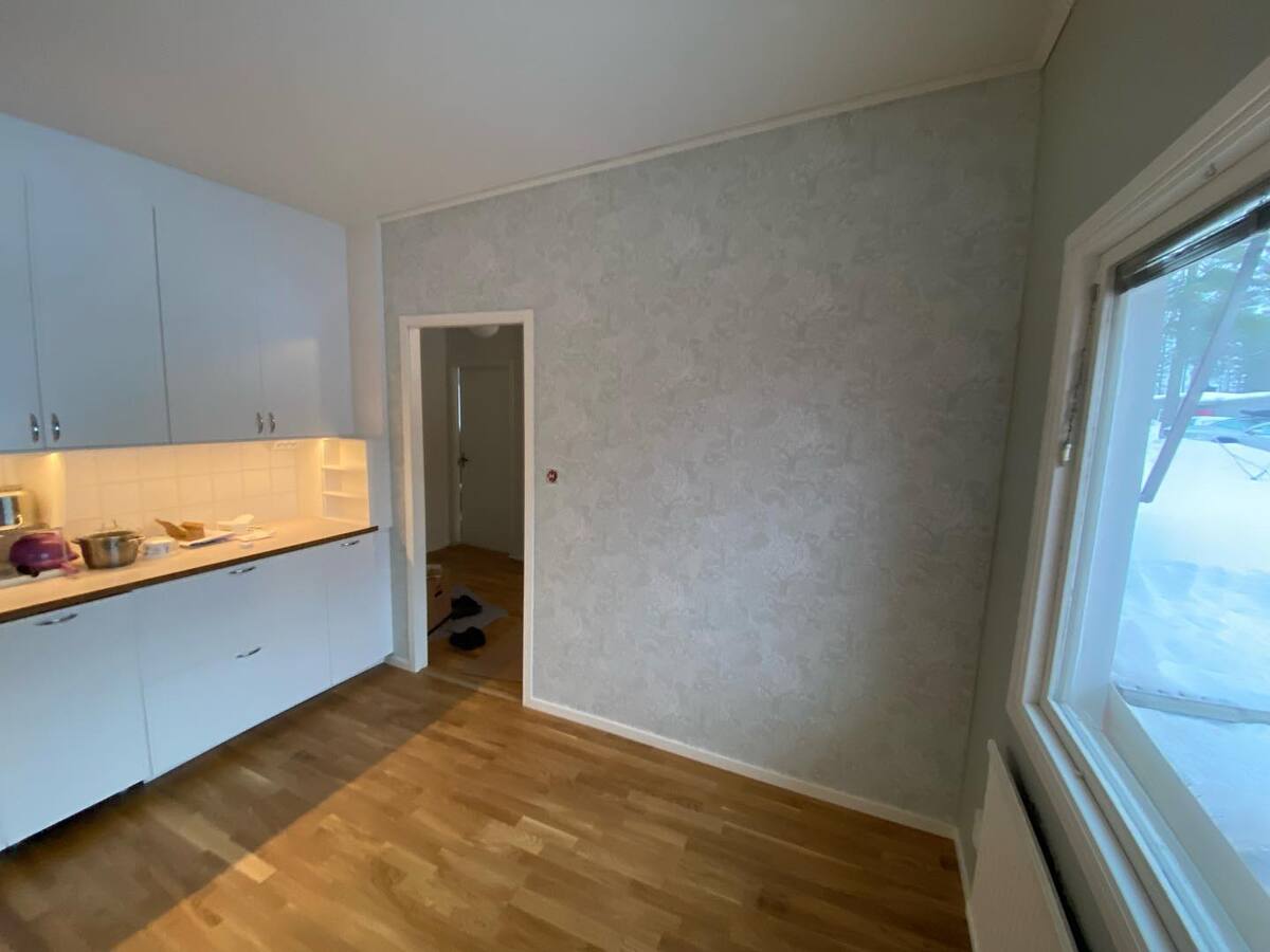 Kök med vita skåp och trägolv, fotat från hörn med utsikt mot fönster och dörröppning.