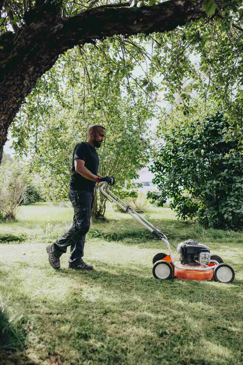 En person går bakom en gräsklippare och klipper gräset i en solig trädgård med träd och buskar. En person går bakom en gräsklippare och klipper gräset i en solig trädgård med träd och buskar.