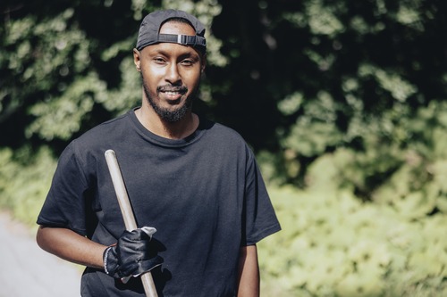 Porträtt av en man med keps och arbetshandskar som håller i ett trädgårdsredskap.