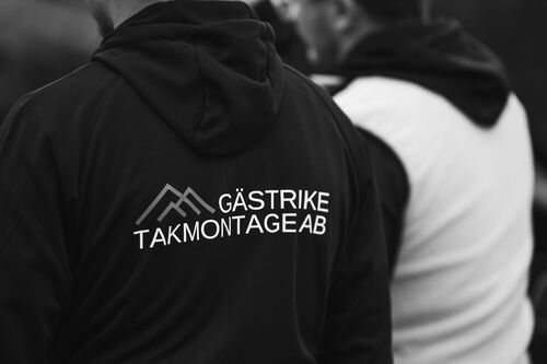Svartvit bild på person i hoodie med Gästrike Takmontage AB-logotyp på ryggen, stående bredvid en kollega i vit väst.