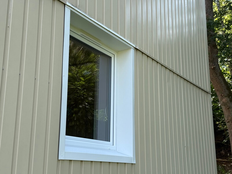 Närbild på nyinstallerat fönster med vit karm i hörnet av ljus träpanelfasad