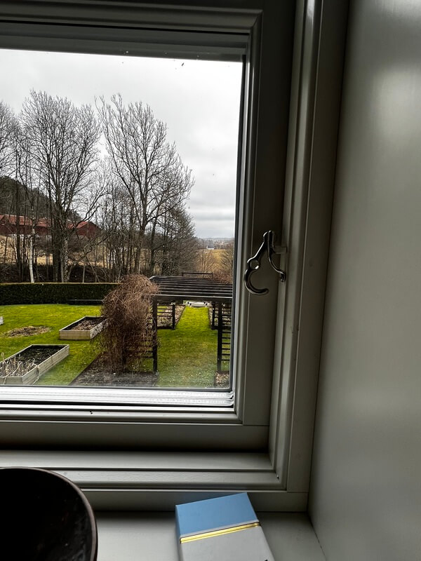 Öppnat sidohängt fönster med mörk karm och utsikt över trädgård med odlingslådor