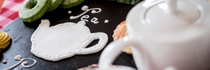Hard white icing in the shape of a teapot with a white ceramic teapot and different biscuits laid around the teapot