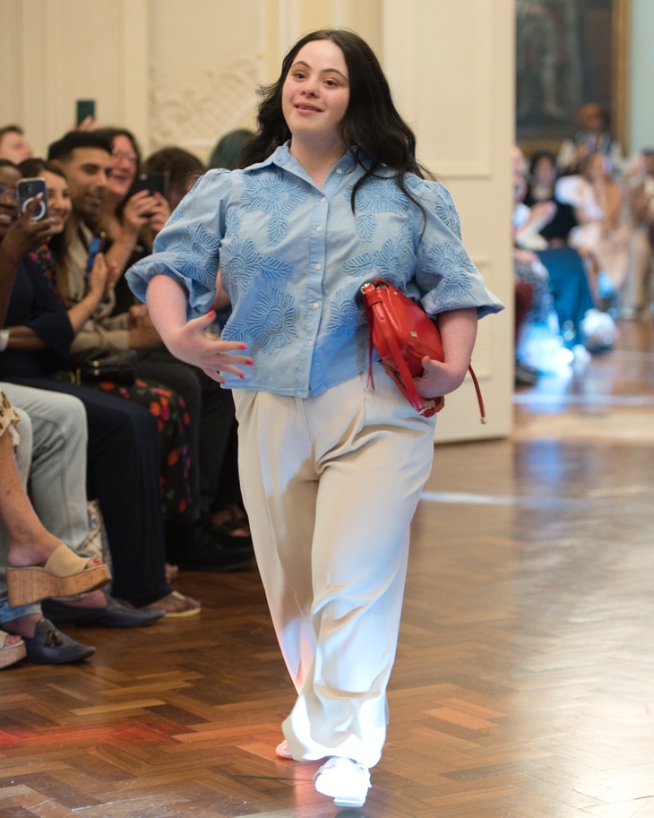 A model wearing a blue shirt, white trousers and red handbag walking down the runway