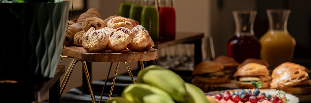 A croissant, strawberry jam spread out on a small round plate with small square-cut pieces of butter and small jam jars laid on the side