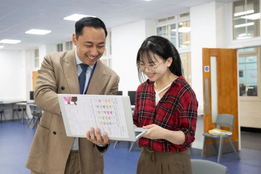 Two staff members are standing in an open classroom, smiling and pointing at a laptop and a colourful chart.