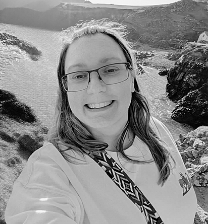 Louise, a white woman with long brown hair and rectangular glasses smiles at the camera in front of a river background