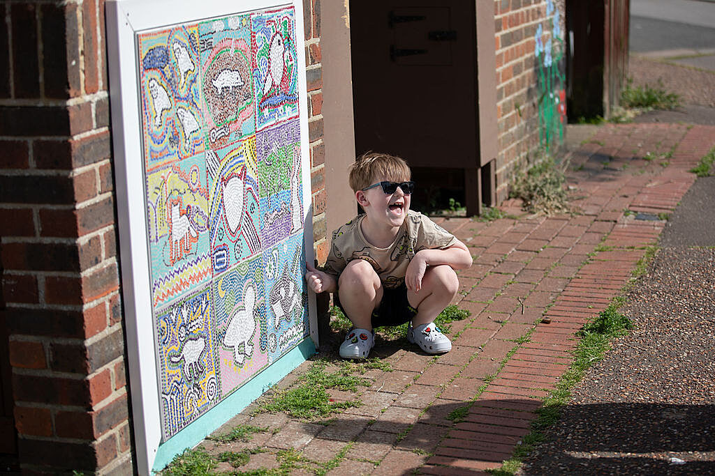 Young boy in sunglasses poses with his thumb up smiling next to a mosaic mural on a wall