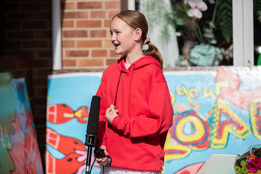 Young girl wearing red hoodie smiling whilst speaking into a microphone