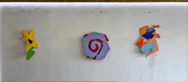 Three abstract, colourful wall sculptures hanging against a gallery wall. They resemble the shape of a flower, spiral, and face