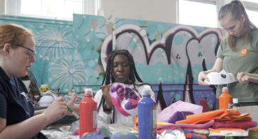 Three young people are sat at a table decorating face masks. They are using collage materials, paint, and fabrics.