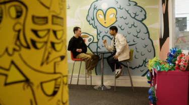 Two young people sit on stools opposite each other chatting