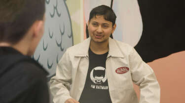 A young man smiles at a peer with a mural in the background