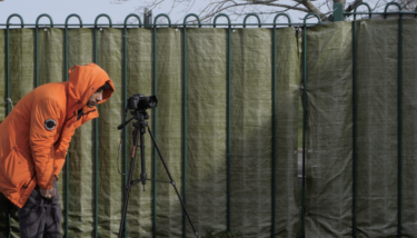 A young person wearing a bright orange coat is looking into a film camera on a tripod next to a tall fence