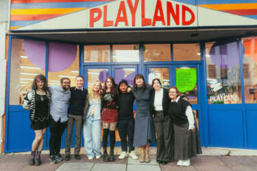 9 young people (under 30) stand outside Playland with the sign in the background - the people are a mix of genders and ethnicities.