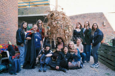 A group of young people standing around a sculpture they created, made from willow and a variety natural materials