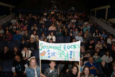 Young people sit in an auditorium with their families and friends. 4 young people stand at the front with a sign that reads 'seaweed rocks'
