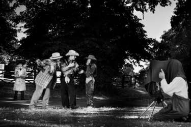 A group of young people dressed as characters in a play are having their photograph taken by another young person with a vintage camera. The image is in black and white to fit the theme of the play.