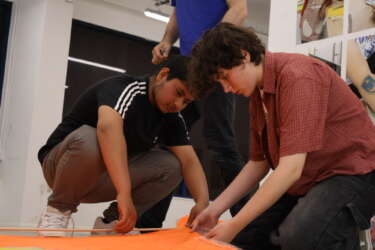 two young people crouched down holding orange fabric and a dowel to make a kite
