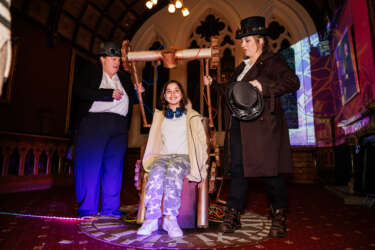 a girl sat on a chair fashioned to look like a Time Machine with a clock face underneath her feet, operated by two people in steam punk style outfits with top hats and welding goggles