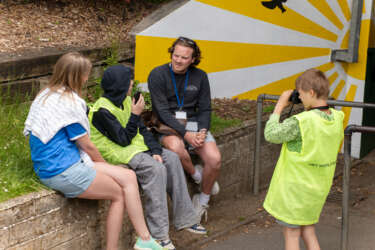 a man being interviewed by a young boy, with another young boy filming and a woman sat listening, all in front of a ray of sun mural