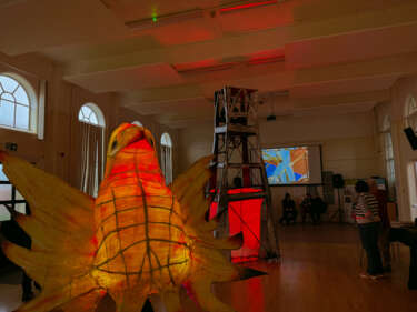 Two large glowing willow paper lanterns. a yellow bird with wings outstretched and an industrial looking tower with mining history influences