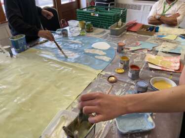 hands visible collaboratively painting a countryside scene onto a large bit of paper