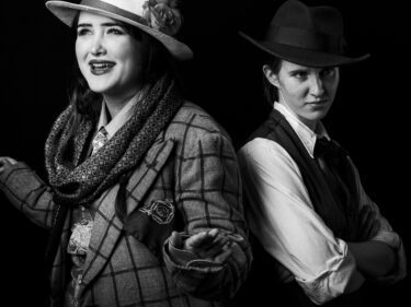 Two young people are dressed in cowboy/western-style clothing for a photoshoot as part of a play they were creating. The young person on the left is smiling with their arms slightly raised, the young person on the right looks intensely at the camera with arms folded.