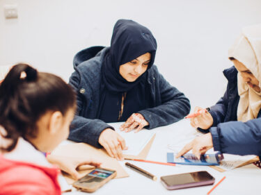 Three young people sat around a table creating poster designs and using their smartphones to search for ideas
