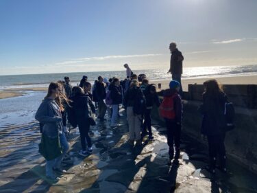 a group of children gather on a beach in the winter sun, facing a man standing on a wall addressing them