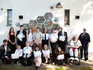 A group of 12 young people stood with an artist and the local town mayor in front of a mosaic mural they helped design and create for a community garden