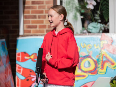 Young girl wearing red hoodie smiling whilst speaking into a microphone