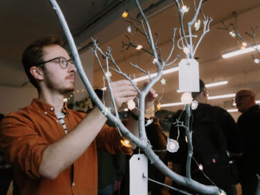 A young man with brown hair wearing an orange button-up shirt and glasses is decorating a tree branch with fairy lights and handwritten notes.
