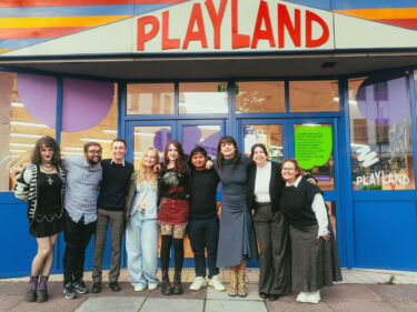 9 young people (under 30) stand outside Playland with the sign in the background - the people are a mix of genders and ethnicities.