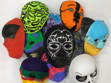 A pile of colourful masks made by young people. Different paints, collage materials, and fabrics have been used to decorate them