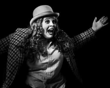 A young person dressed in vintage clothing as part of a photoshoot for a play they were creating. They are wearing a bowler had and tweed jacket. They have their hands raised in excitement and mouth open as if they are shouting.