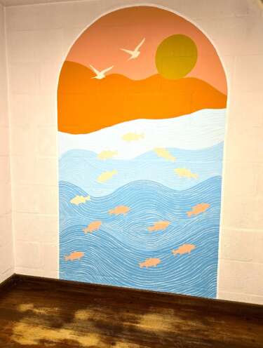 An arch shaped mural seascape showing waves in a gradient of blues with orange fish, and an orange sunset sky with birds flying, painted onto a plain wall