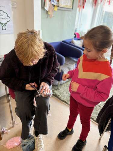 A woman and a child sit together making pompoms. The woman is cutting the handmade pompom and the child is looking at it.