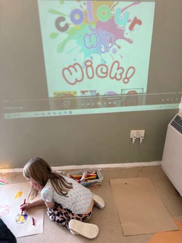 a young girl sits on the floor drawing on a piece of paper. Projected on the wall behind her is the poster for 'Colour up Wick'.