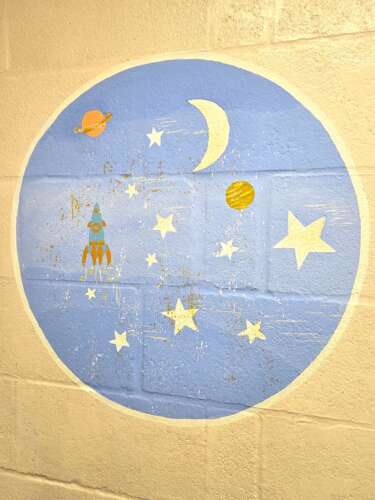 A circular mural with blue background and pastel moon, stars, planets, and rocket, painted onto a plain wall