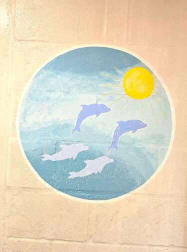 A circular mural in pastel colours of dolphins swimming and jumping with a sun, painted onto a plain wall