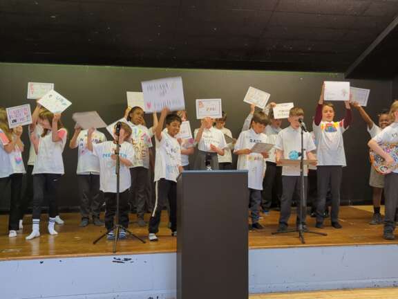 Year 5 pupils on stage wearing decorated t-shirts and holding signs with messages on them, in performance