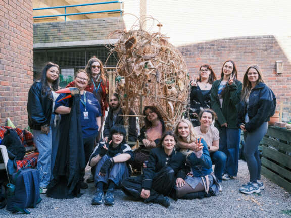A group of young people standing around a sculpture they created, made from willow and a variety natural materials