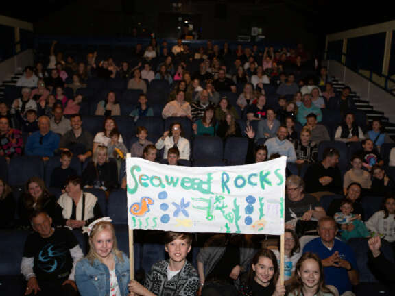 Young people sit in an auditorium with their families and friends. 4 young people stand at the front with a sign that reads 'seaweed rocks'