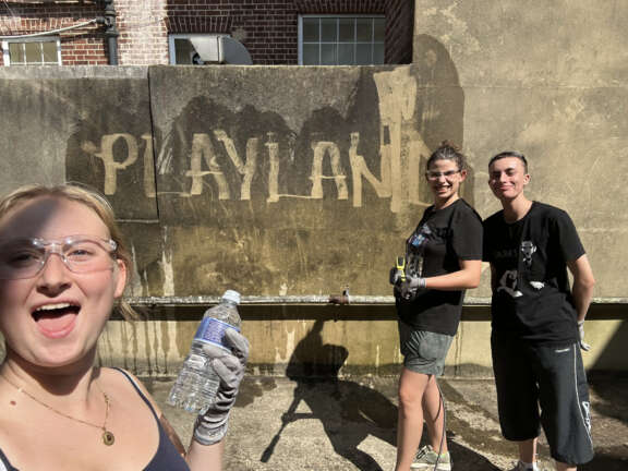 3 people work on Playland garden and on the wall they have used a jet wash to write 'Playland' on a muddy wall.