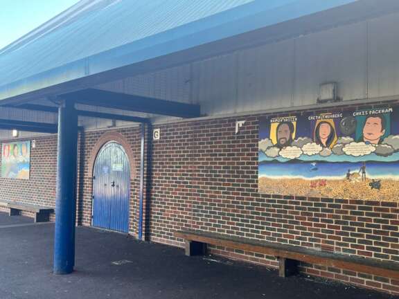 painted mural panel affixed to brick school wall. mural contains simplified painted portraits of hamza yassin, greta thunburg, and chris Packham. underneath the portraits there are quotes in speech bubbles that aren't readable from this image, and a painted beach scene